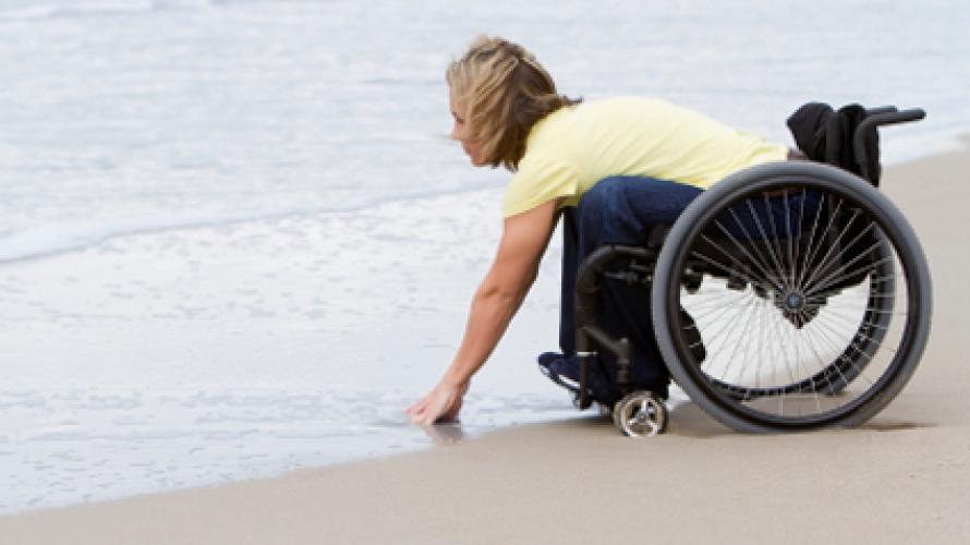 Persona en silla de ruedas en la playa, inclinándose para tocar el agua